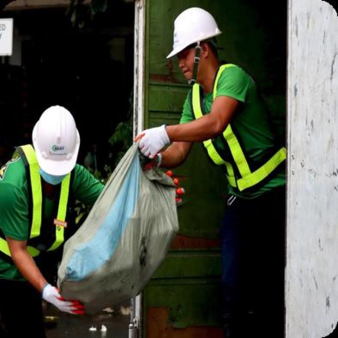 Two crew members in hard hats and high-visibility vests lifting a bagged load of recyclables out of a storeroom