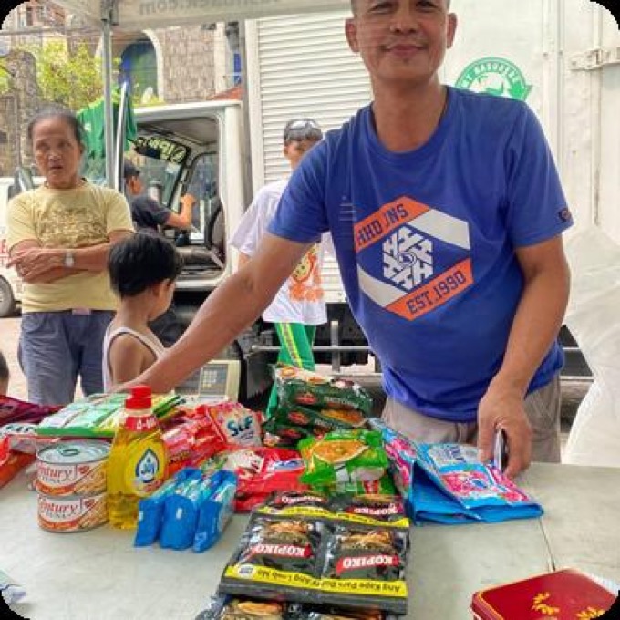 A resident smiling behind a table of groceries — canned goods, cooking oil and noodles — claimed with Environmental Points