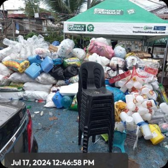 Sacks of pre-sorted plastics and containers piled beside a green Trash to Cashback pop-up tent on a community collection day